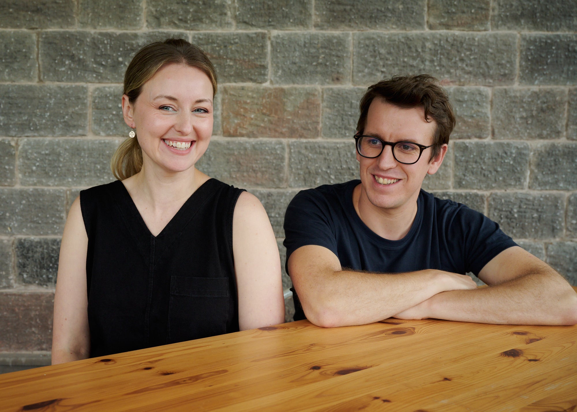 Photograph of Joe and Gemma, the founders of Unspun, smiling and seated at a wooden table. Joe wears a dark blue t-shirt and glasses, and Gemma is in a black sleeveless top. They are positioned against a backdrop of a textured brick wall. This image is part of the 'About Unspun' page, introducing the faces behind the brand.