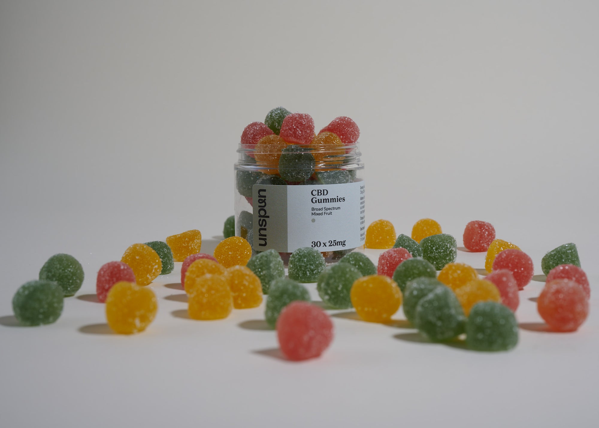 A scattering of colorful CBD gummies surrounds an open jar labeled 'Unspun CBD Gummies, Broad Spectrum, 30 x 25mg,' evoking a sense of plentifulness and variety, reflecting the 'gummies of abundance' theme of the product photo shoot.