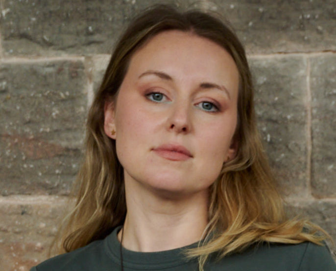 A close-up portrait of Gemma, one of Unspun's founders, wearing a casual olive t-shirt against a brick wall. Her expression is thoughtfully aloof, adding a comical touch that hints at the brand's approachable and unique character