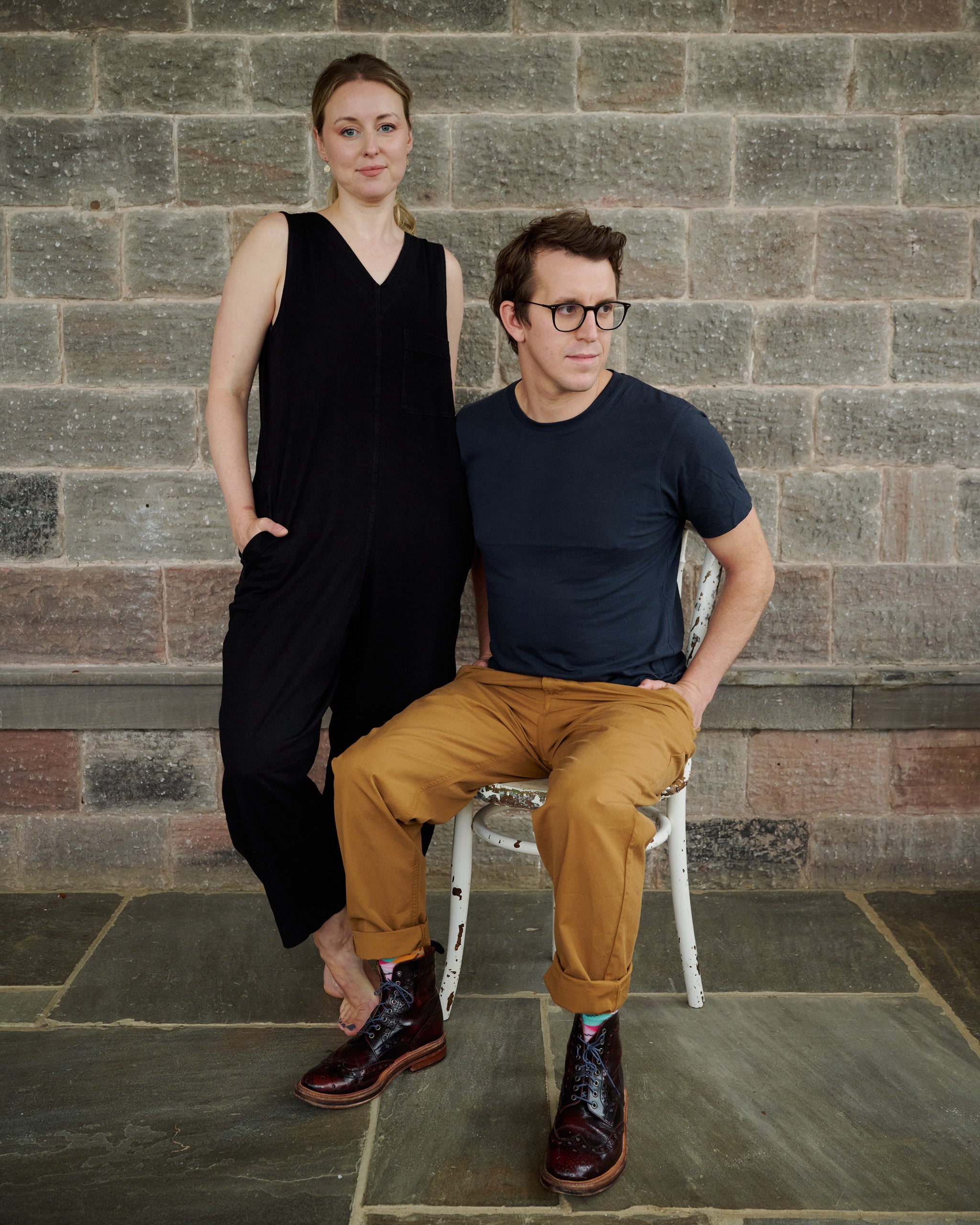 Portrait of Unspun's founders, Gemma and Joe, posing against a brick wall backdrop. Gemma stands with one hand on her hip, wearing a black sleeveless dress, while Joe sits on a white chair, dressed in a blue t-shirt and tan pants, with distinctively patterned socks and brown leather shoes.